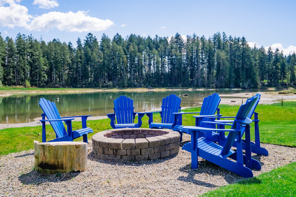 A fire pit surrounded by five blue Adirondack chairs is situated on pebbled ground. The serene water reflects the trees in the background, creating a tranquil outdoor space, ideal for relaxation and gatherings.