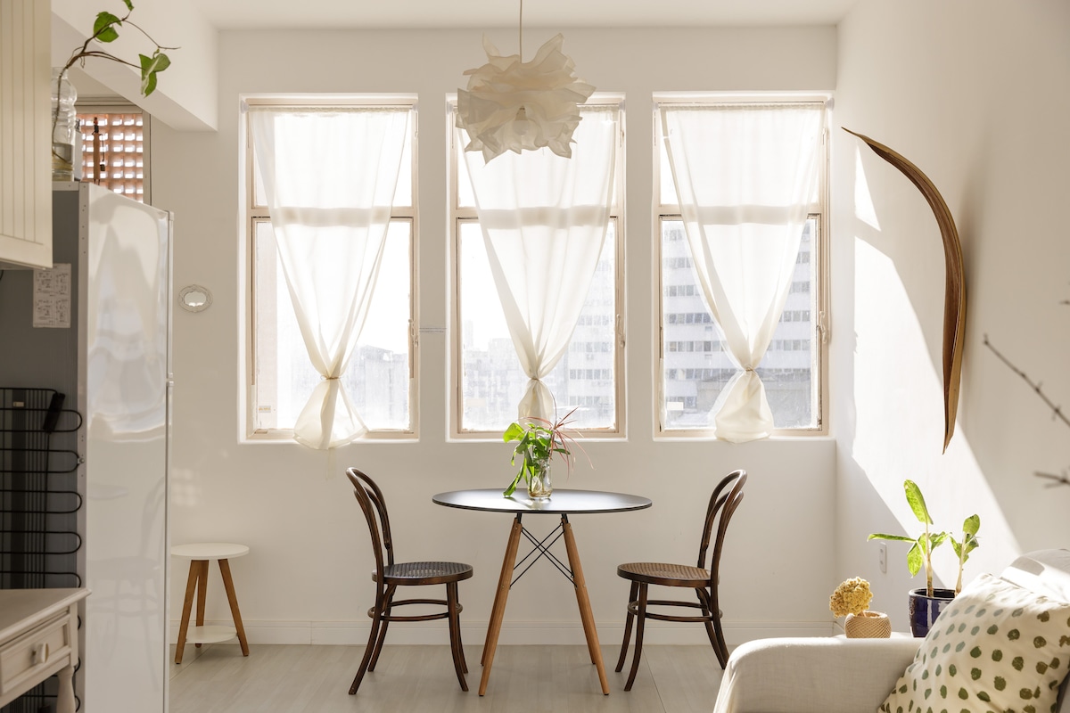 A bright dining area is showcased with a round table surrounded by two wooden chairs. Natural light streams through three large windows, adorned with sheer curtains. A small potted plant adds a touch of greenery to the inviting space.