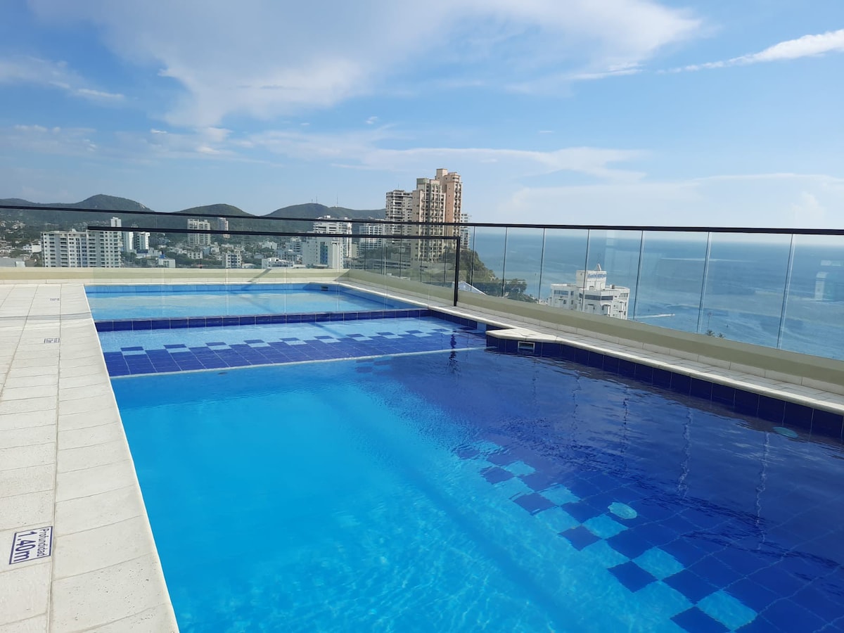 An expansive infinity pool is located on a rooftop terrace, offering panoramic views of the surrounding landscape and ocean. The water shimmers in a bright blue hue, with reflections of the clear sky visible. Nearby, modern buildings are seen against a backdrop of distant hills.