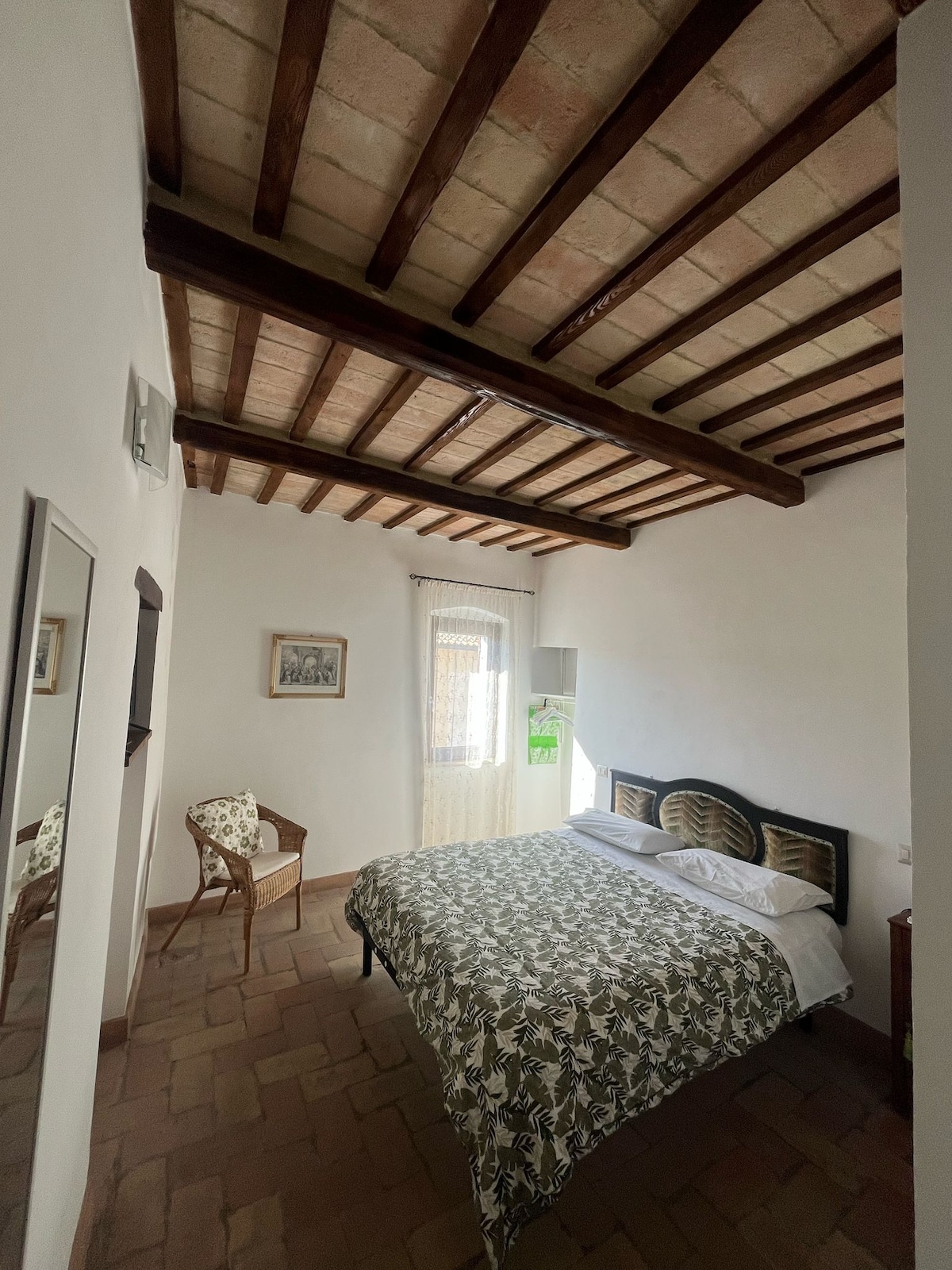 A spacious bedroom features a comfortable bed adorned with a botanical print duvet. Exposed wooden beams run along the ceiling, while a window allows natural light to fill the room. A mirror and a charming chair complement the rustic decor.