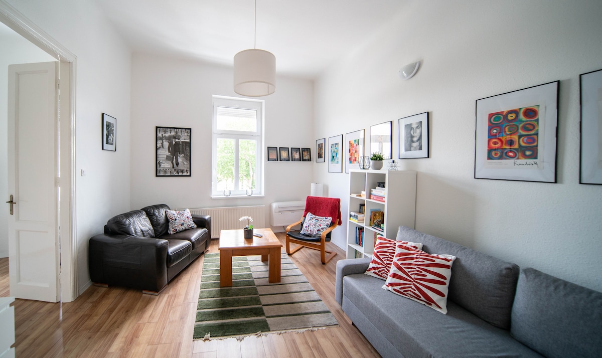 A bright living area features two comfortable sofas, a wooden coffee table, and decorative throw pillows. Wall art in various styles and sizes is displayed around the room. Natural light enters through a window, enhancing the spacious feel of the minimalist design.