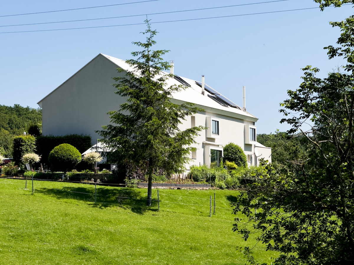 The exterior of the house is visible, showcasing a modern two-story structure with a light-colored facade and large windows. Solar panels are mounted on the roof, while a well-maintained garden with various shrubs and trees surrounds the property, creating a peaceful natural setting.