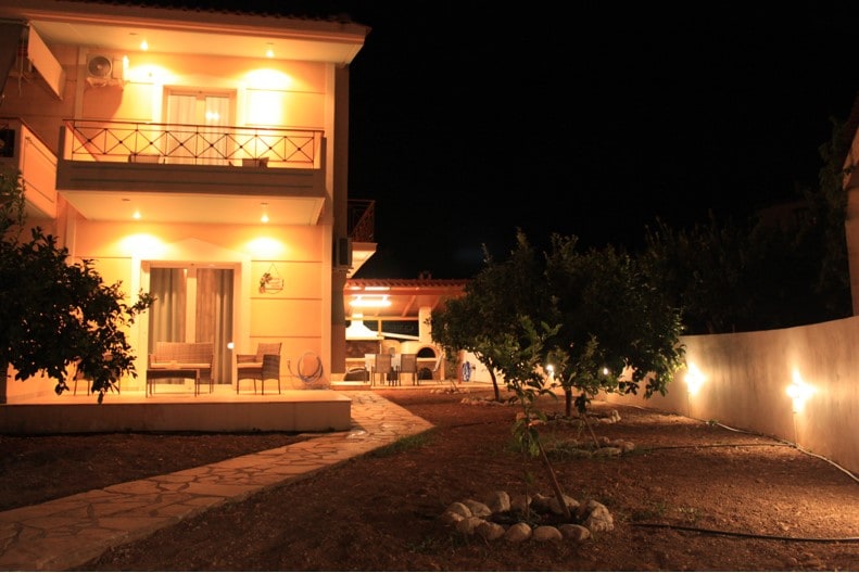 The exterior of the house is illuminated warmly at night, showcasing two levels and a stone pathway leading through the garden. Soft lighting highlights trees and shrubs, creating a cozy ambiance around the outdoor seating area beneath the covered patio.