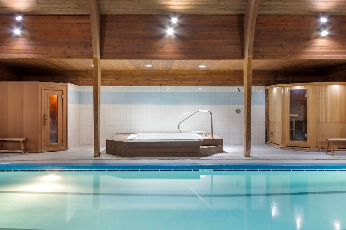 An indoor pool area is featured, illuminated by recessed lighting. A wooden sauna and a whirlpool jet tub are positioned alongside the pool, creating a relaxing environment. The warm wood tones of the ceiling and walls add to the tranquil atmosphere.