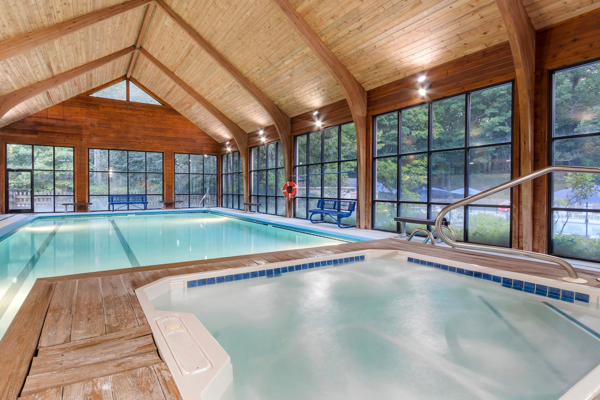 An indoor pool area features high wooden ceilings with large windows allowing natural light to flood the space. A hot tub is situated beside the pool, offering a relaxing atmosphere, while rows of benches line the perimeter for seating.
