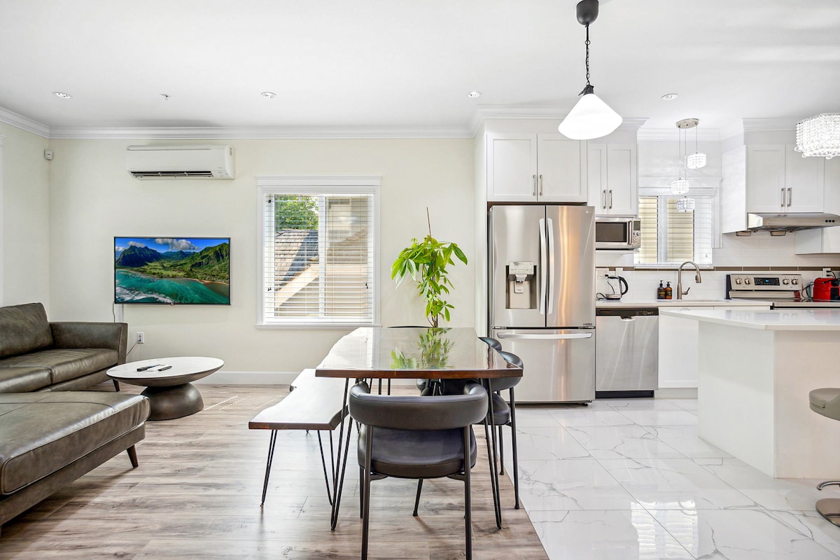 An open-concept living area features a modern kitchen with stainless steel appliances and a dining table surrounded by stylish chairs. A comfortable sofa is positioned next to a large wall-mounted TV, with natural light streaming in through the windows.