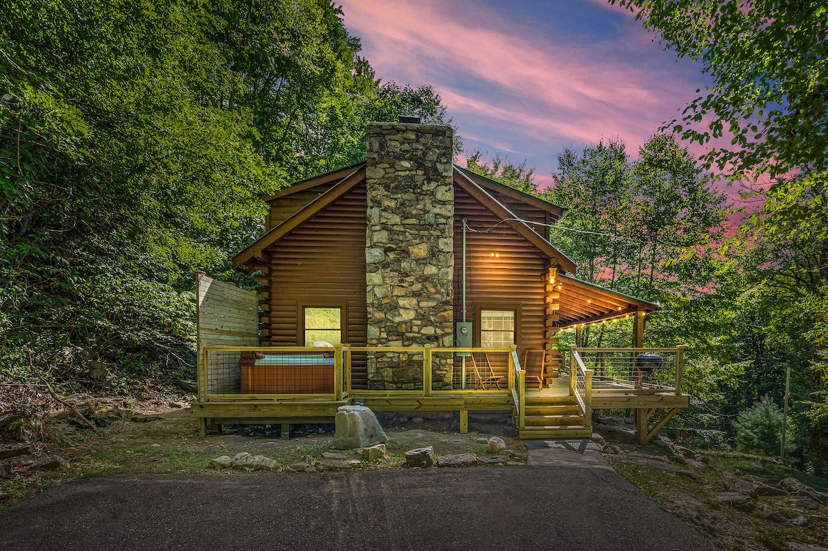 A cozy log cabin features a stone chimney and wooden deck, surrounded by lush, green foliage. A hot tub is situated on the deck, with warm evening light illuminating the space, creating a welcoming atmosphere for relaxation.