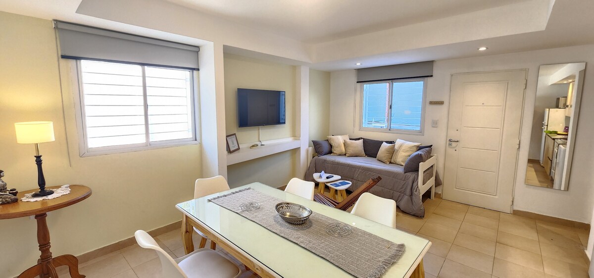 A light-filled living area features a neutral palette with a dining table set for six. A flat-screen TV is mounted on the wall opposite a spacious sofa, complemented by large windows allowing natural light. The open layout connects seamlessly to the kitchen.