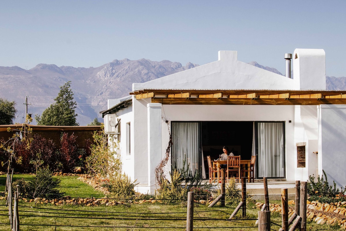 A self-catering cottage features a white exterior with large glass doors opening onto a private patio. The patio includes a wooden dining set surrounded by greenery, with mountains visible in the background under a clear blue sky.