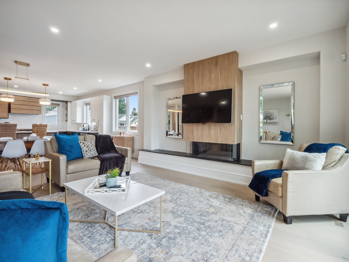 A modern living space features a sleek television mounted above a cozy fireplace, flanked by mirrors reflecting the natural light. Two comfortable sofas with blue accents surround a central coffee table atop a detailed area rug, creating an inviting atmosphere for relaxation or socializing.