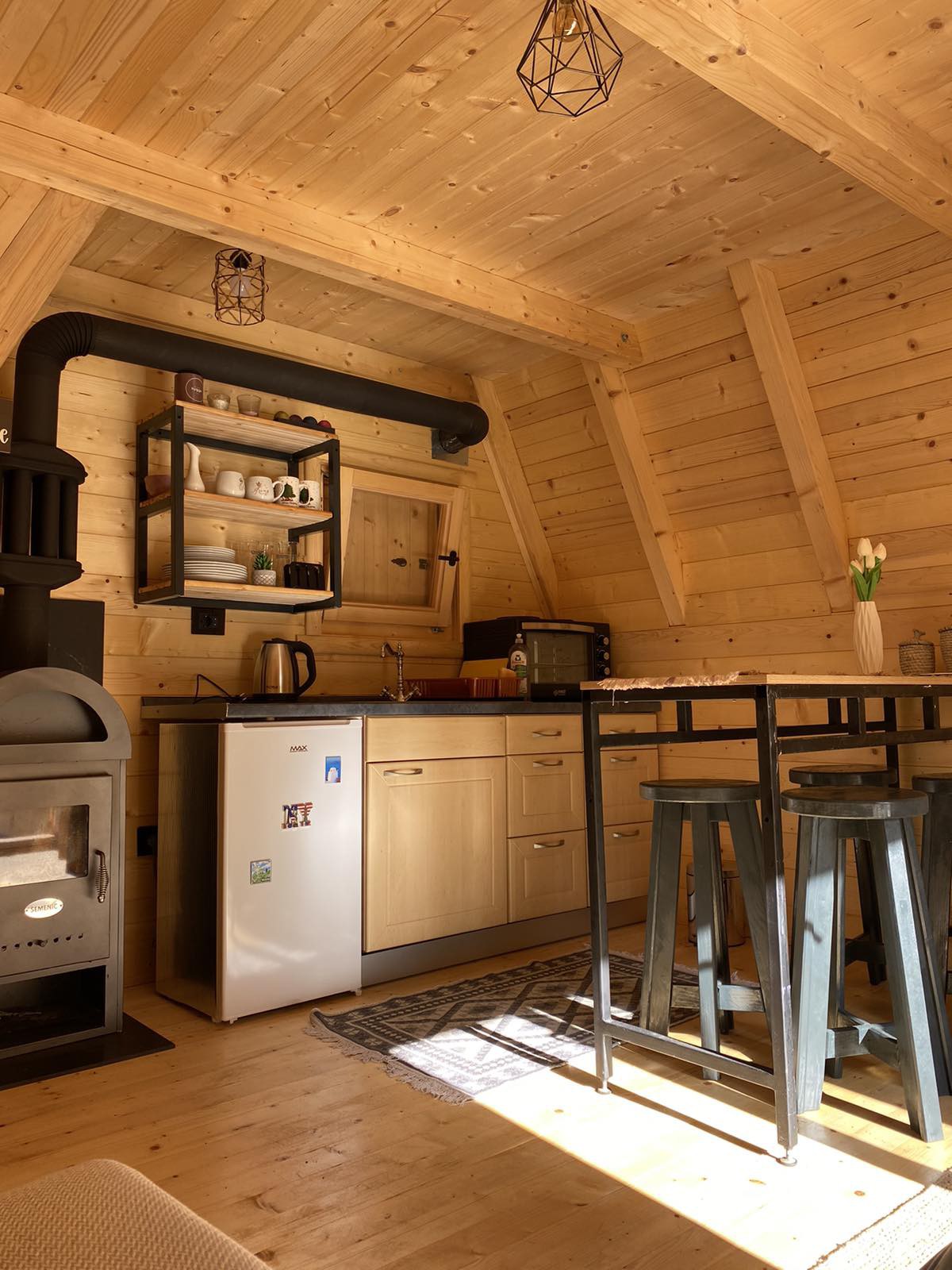 A compact kitchen area is showcased with wooden walls and ceiling, illuminated by natural light. Modern appliances including a refrigerator and stove are present, alongside open shelving for dishware. A dining table with high stools allows for cozy meals, enhancing the space's functionality.