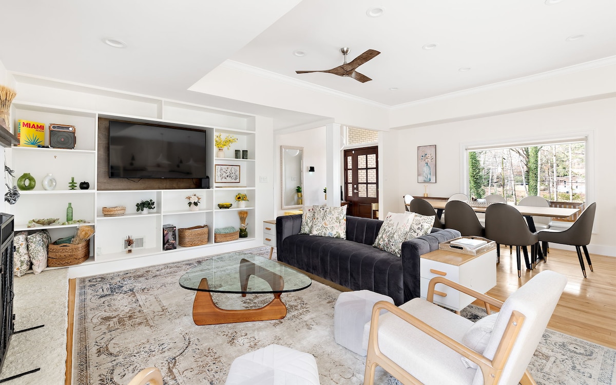 The spacious living room features a dark sectional sofa, a coffee table, and a plush area rug. A large TV is mounted above a stylish console, while natural light fills the room through a wide window overlooking the forest. Dining chairs are placed nearby.