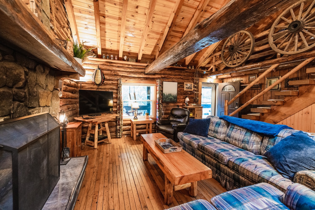 A spacious living area features a stone fireplace and a large wooden table. Comfortable seating arrangements include a plaid couch and a leather armchair. Natural wood tones define the cabin's interior, with a set of stairs leading to an upper loft area. Large windows invite natural light.