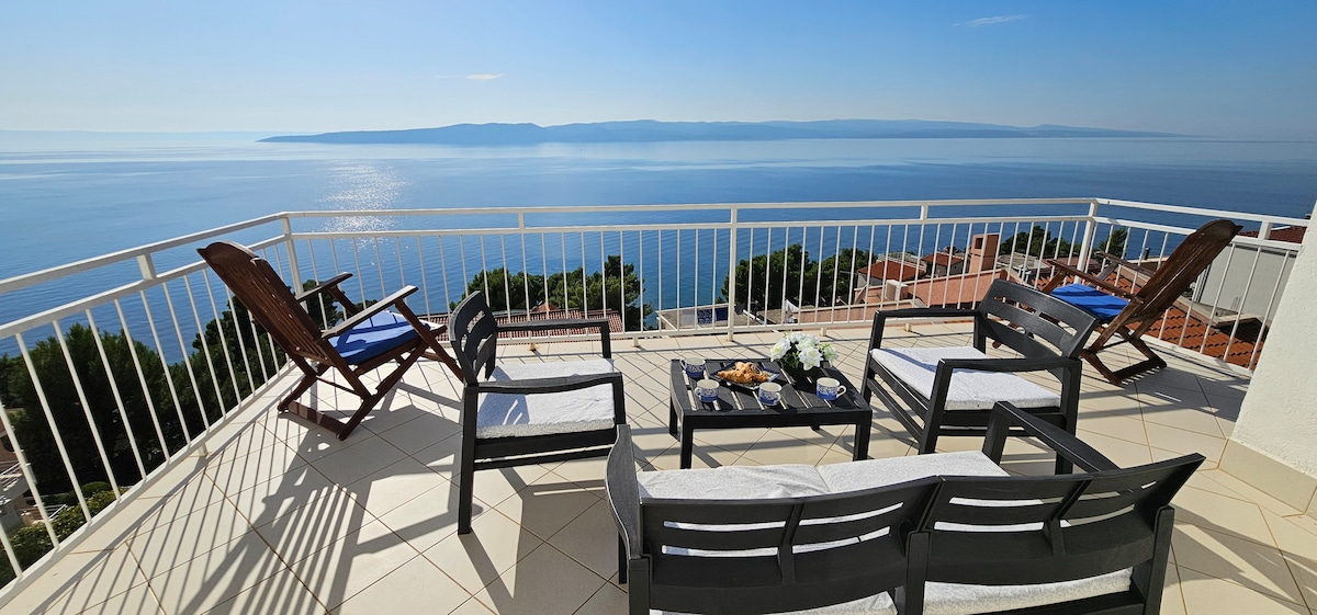 A spacious terrace showcases comfortable seating arrangements, including reclining chairs and a low table. Breathtaking views of the Adriatic Sea create a stunning backdrop, with clear skies reflecting sunlight on the water's surface, inviting relaxation and enjoyment of the surrounding scenery.