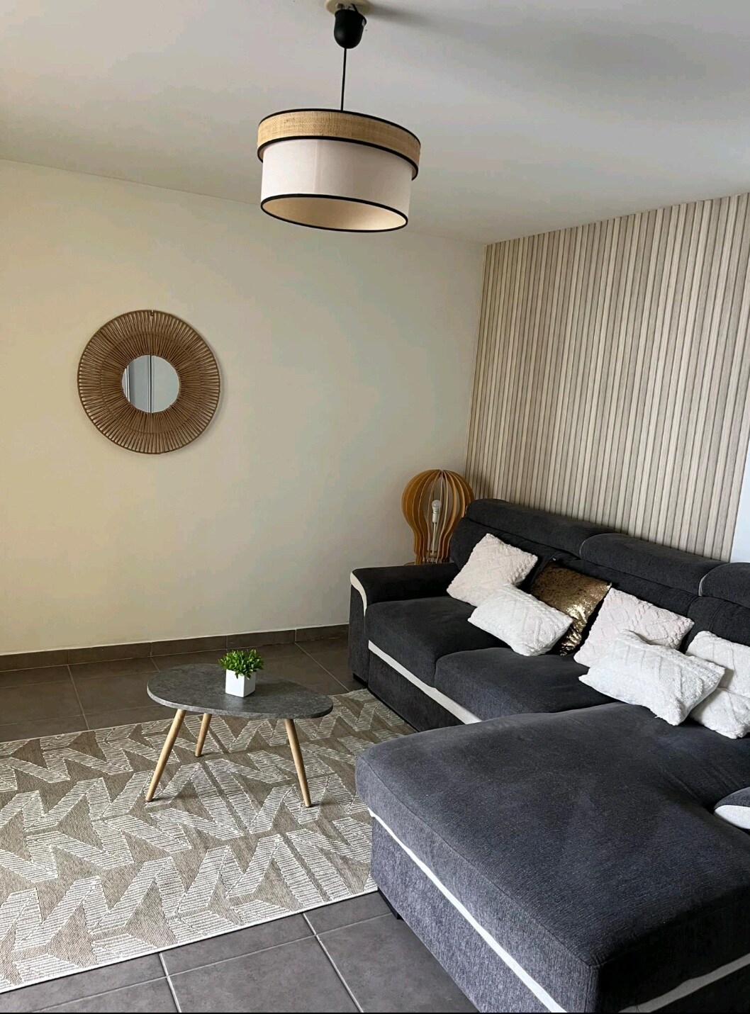 A comfortable living area is shown, featuring a dark sectional sofa with various textured cushions. A round coffee table sits atop a patterned rug, while a woven mirror hangs on the wall, reflecting natural light. The space is designed for relaxation and socializing.