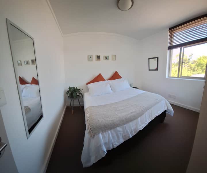 Clean and cozy Bedroom 2 with a queen bed, fresh linen, bedside table and lamp, a lush plant, large windows for natural light, and built-in cupboards for extra storage.