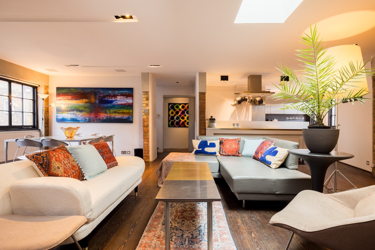 A spacious living area features a blend of mid-century and modern Italian furniture. A large colorful wall art piece adds a vibrant touch, while a green plant provides a natural element. Soft lighting illuminates the seating area, enhancing the inviting atmosphere.