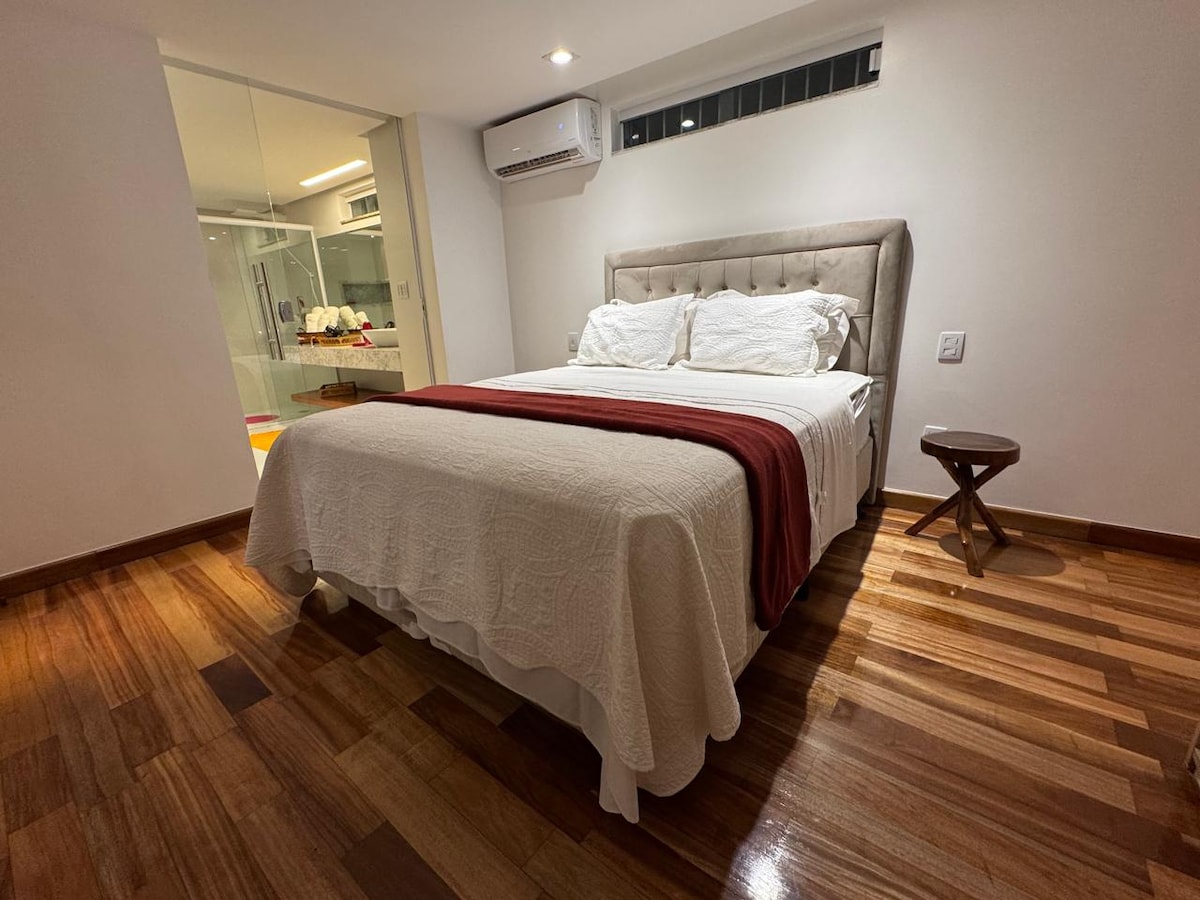 A modern bedroom features a spacious bed dressed in white linens with a maroon accent. Natural wood flooring complements the soft tones. A compact side table is positioned beside the bed, and large windows allow for ample light, creating a bright and airy atmosphere.