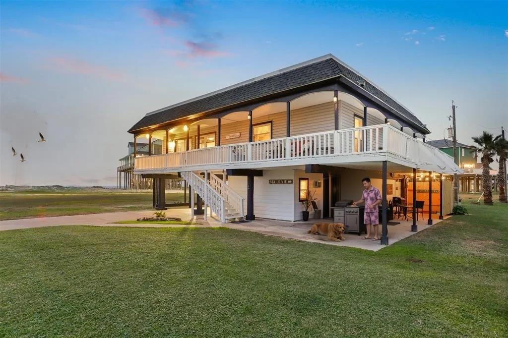 A two-story coastal home is presented with a wrap-around deck, surrounded by a spacious lawn. A person and a dog are seen enjoying the outdoor area, while the setting sun casts a warm glow on the exterior.
