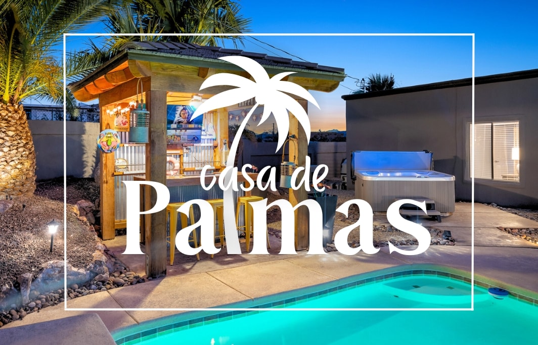 An outdoor space features a covered bar area with seating, surrounded by palm trees. A jacuzzi is situated nearby, with a turquoise pool reflecting evening light. The fencing outlines a cozy atmosphere perfect for relaxation or gatherings.