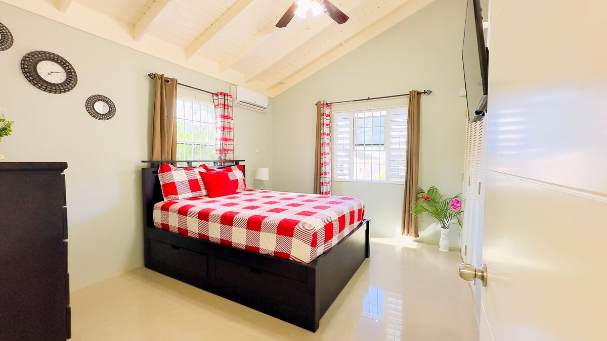 A cozy bedroom features a spacious bed adorned with a red and white checkered blanket. Light enters through a large window, framed by sheer curtains. A dresser stands nearby, complemented by wall decor including circular mirrors, adding a touch of charm to the serene space.