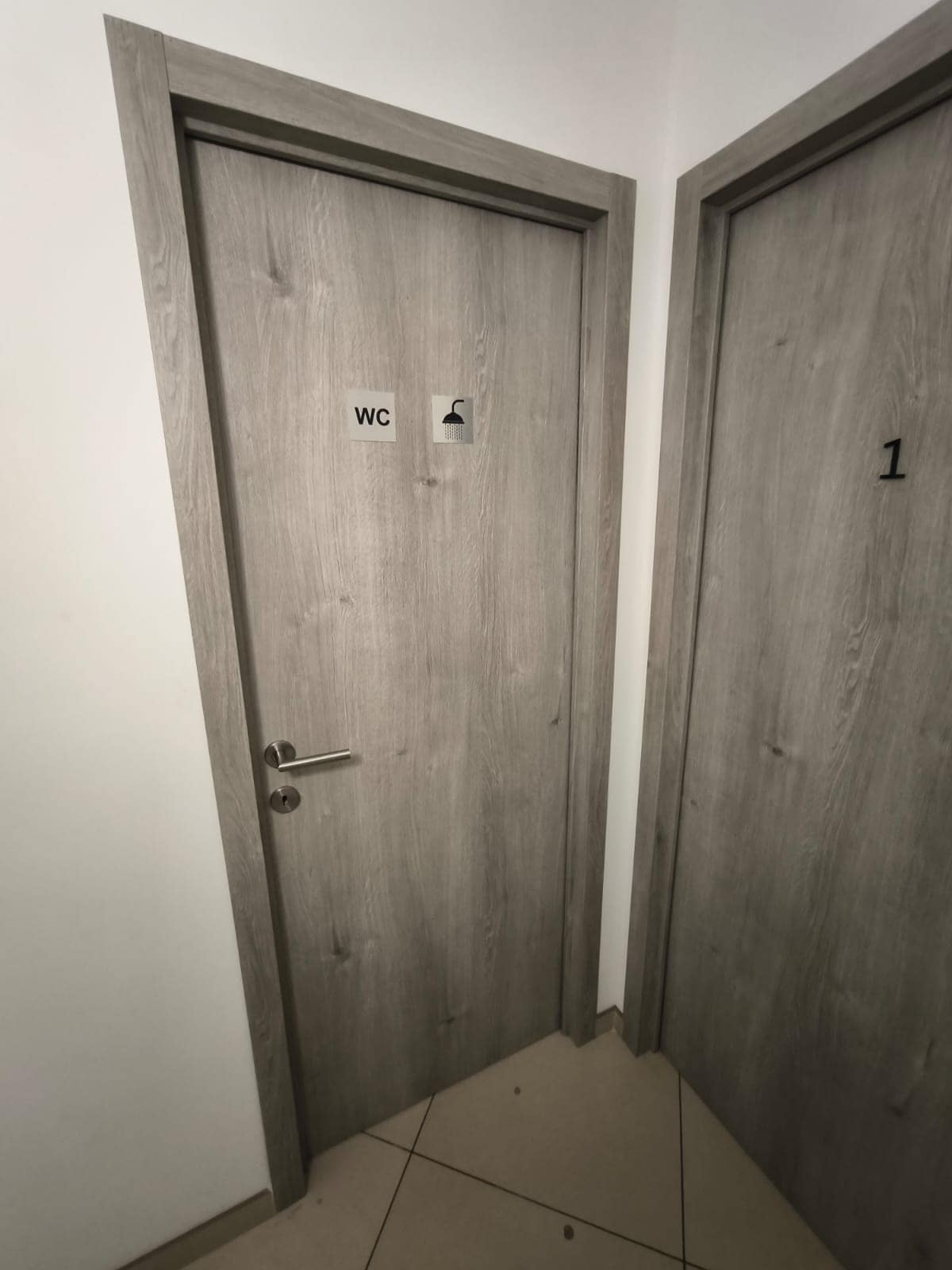 Two wooden doors are visible, labeled with simple signs for identification. One door indicates access to a restroom, while the other is marked with a number. The walls are finished in light tones, and the flooring consists of neutral tiles.