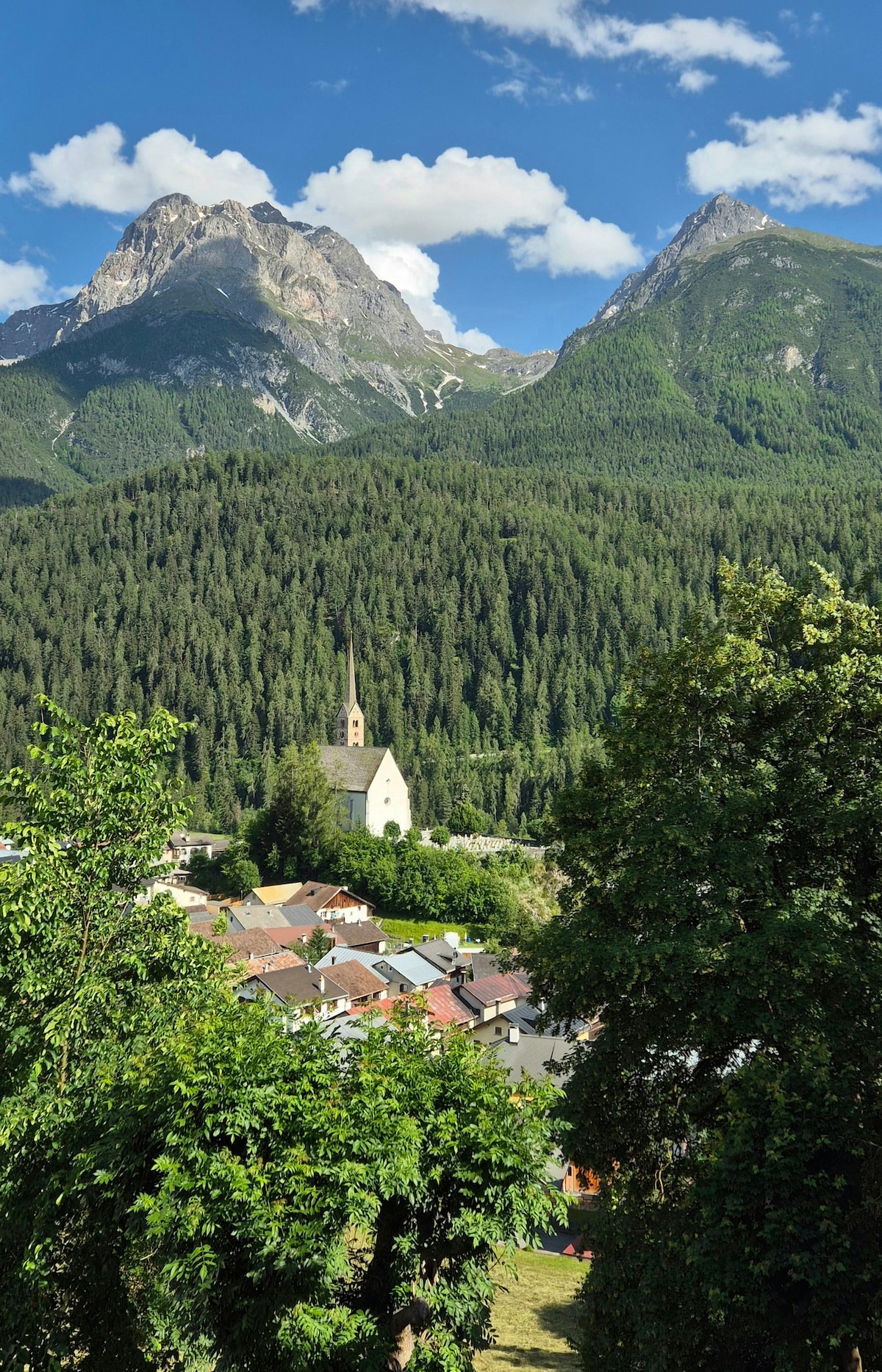 A scenic view captures the surrounding Lower Engadine mountains towering over a quaint village, with a prominent church steeple amidst the greenery. Lush trees frame the foreground, while a cluster of traditional houses can be seen nestled within the valley.