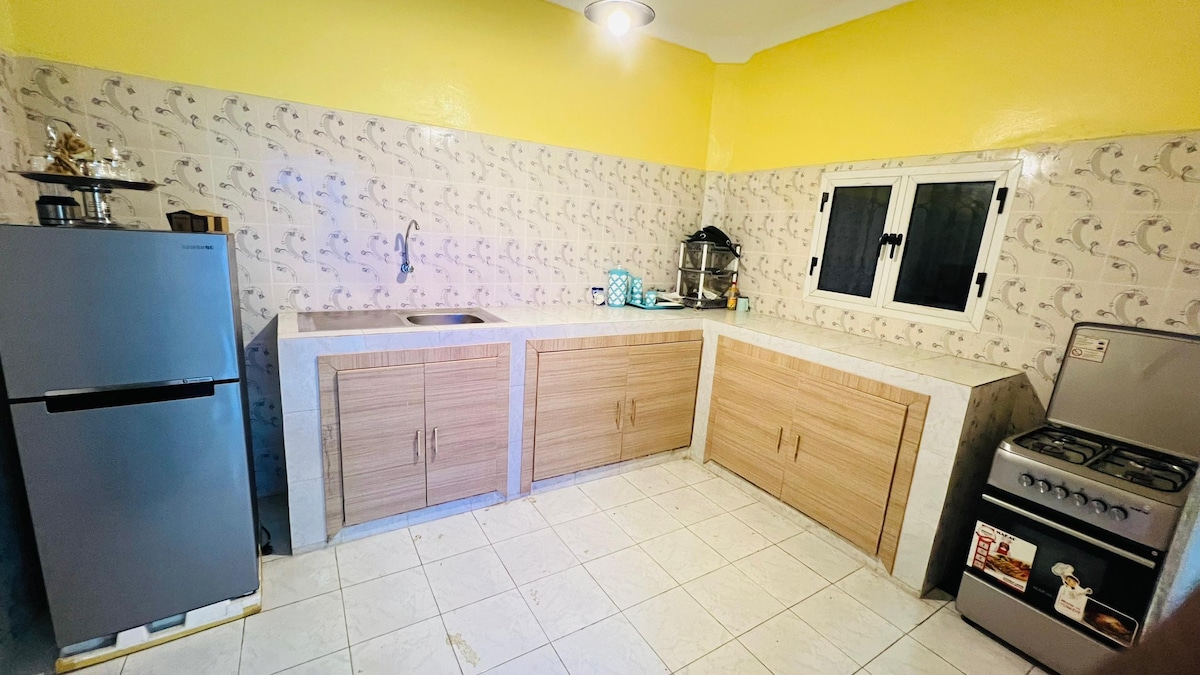 The kitchen features a spacious layout with light-colored cabinetry and countertops. A stainless steel refrigerator and a gas stove are visible, along with a sink and various kitchen items neatly arranged. Bright yellow walls enhance the inviting atmosphere of the cooking space.