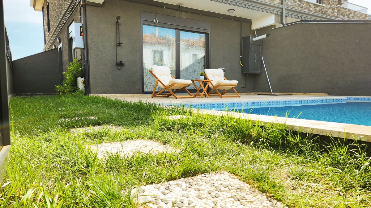 A tranquil outdoor space is highlighted, featuring a refreshing swimming pool beside a neatly manicured lawn. Two comfortable chairs are positioned on a stone pathway, inviting relaxation in the sun. The surrounding walls ensure privacy and a peaceful atmosphere.