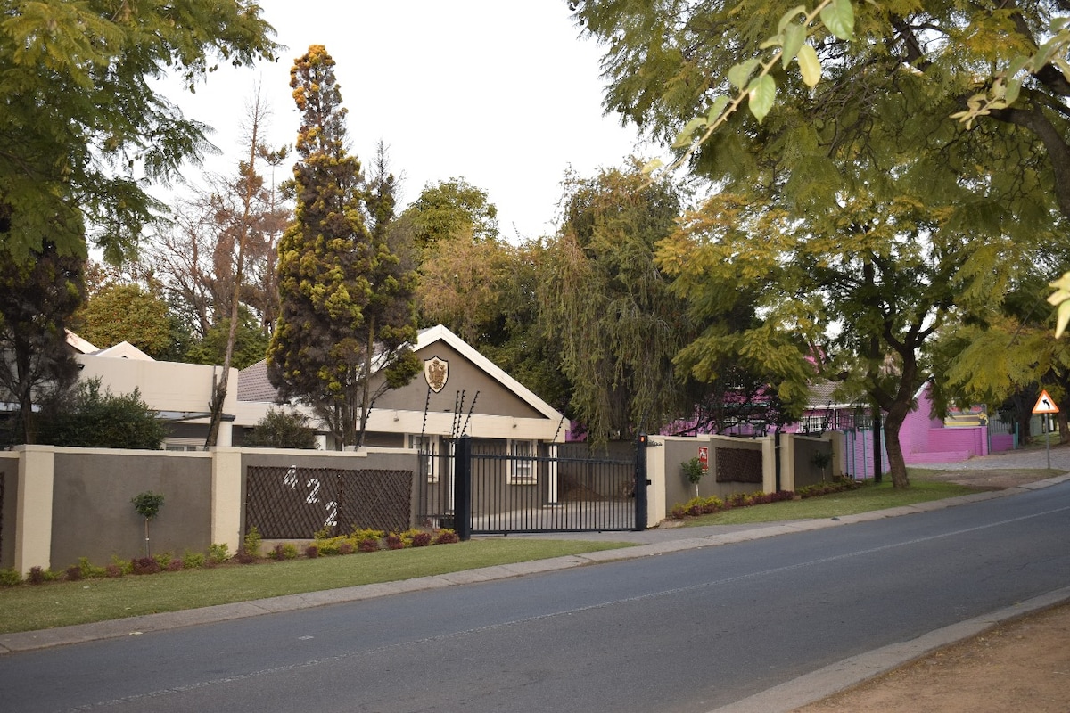 The exterior of the guest house is visible, featuring a gated entrance framed by lush greenery and landscaped flower beds. The building showcases a pitched roof and neutral-colored walls, set against a serene street scene lined with trees. A welcoming atmosphere is evident.