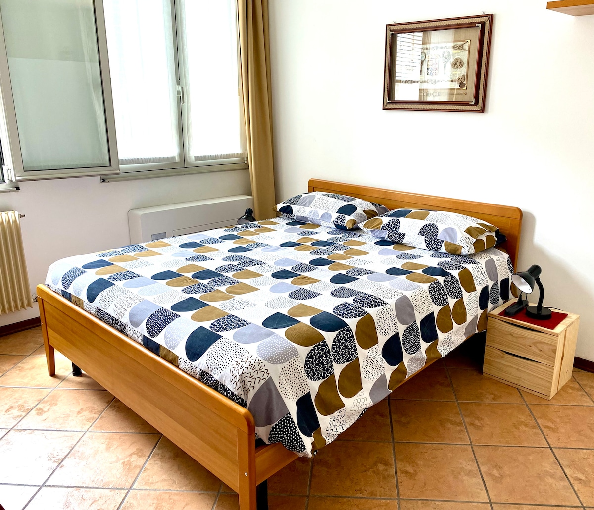 A well-lit sleeping area features a wooden bed with a patterned duvet in shades of blue, yellow, and white. A framed artwork is mounted on the wall above the bed, while large windows allow natural light to fill the room.
