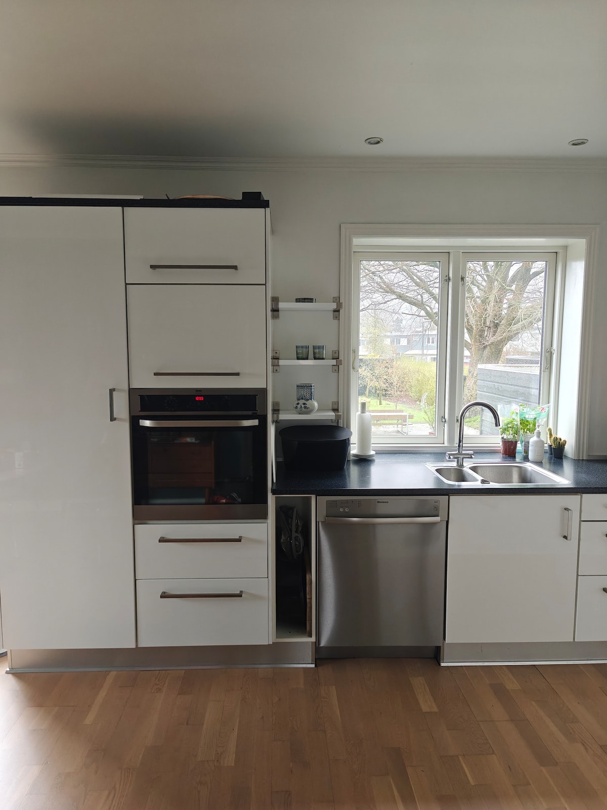 A modern kitchen setup is presented, featuring sleek white cabinetry and stainless-steel appliances. A large window above the sink allows natural light to illuminate the space. Wooden flooring adds warmth, while a few small plants are arranged on the countertop, enhancing the overall freshness of the area.