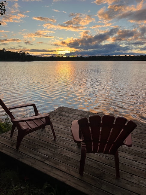 Two red Adirondack chairs are positioned on a wooden deck overlooking Lower Post Lake, where a sunset creates a warm glow on the water's surface. Soft clouds hover in the sky, reflecting shades of orange and purple as day transitions to evening.
