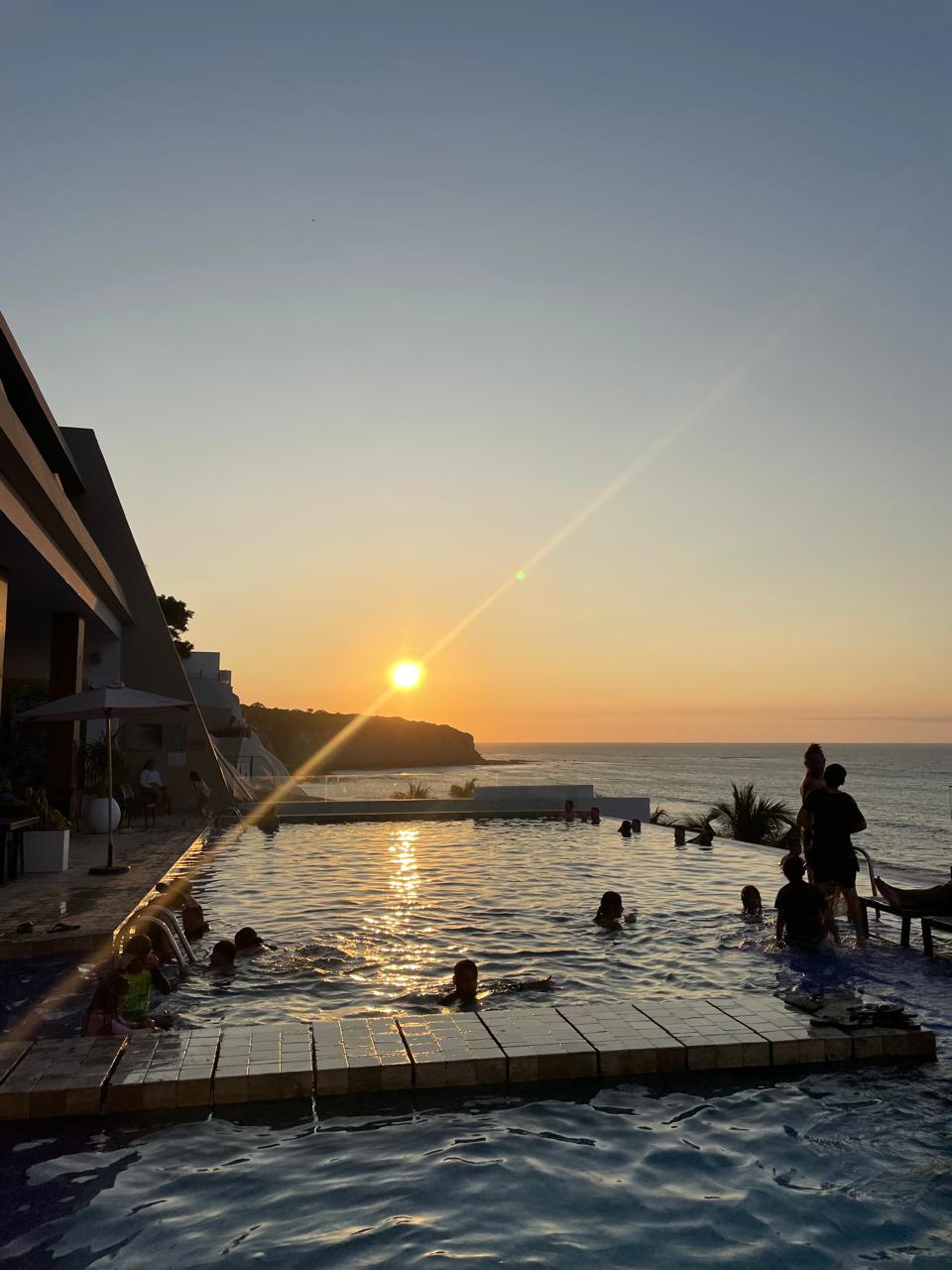An infinity pool extends into the ocean, reflecting the vibrant colors of the sunset. Silhouettes of guests can be seen enjoying the water, with the sun descending towards the horizon. Lush greenery is visible in the background, adding a natural element to the serene scene.