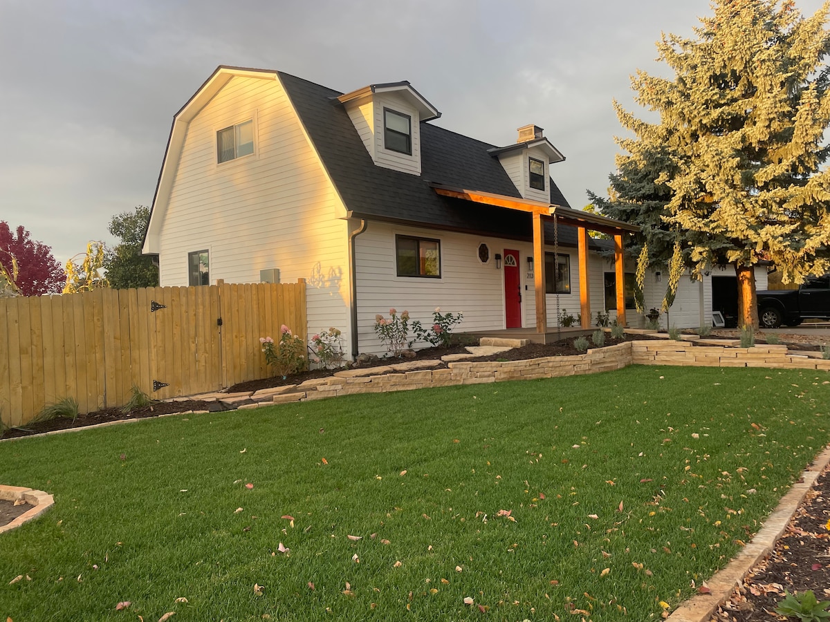 The exterior of the house is showcased, featuring a two-story design with a welcoming red door. A wooden porch is supported by beams, while a manicured lawn and flower beds are visible. A wooden fence encloses the property, with trees partially in view.