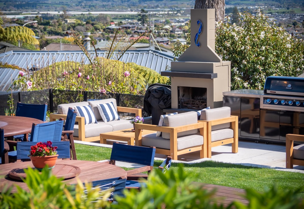 A well-designed outdoor space features modern furniture with grey cushions arranged around a fire pit, complemented by a barbecue grill nearby. A pot of red flowers adds a touch of color, with a scenic view of the surrounding hills and valley in the background.