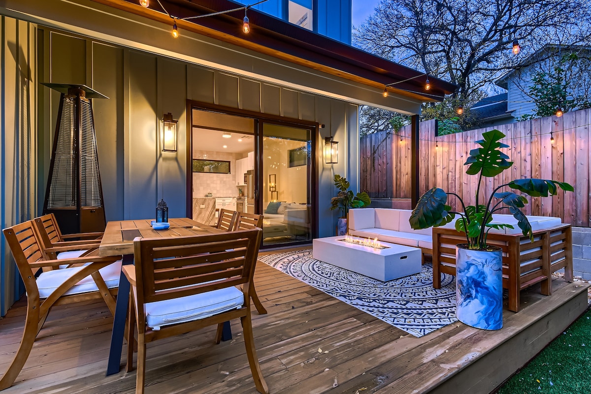 A spacious patio area features a dining table with wooden slatted chairs and a cozy seating arrangement around a fire pit. Twinkling bistro lights hang above, enhancing the warm ambiance. Lush plants are positioned around the space, adding a touch of greenery to the modern design.