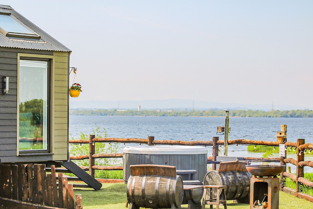 A glamping lodge is positioned near the water's edge, featuring a private hot tub and a fire pit area. Rustic wooden barrels and seating provide a cozy outdoor space. The expansive view of Lough Neagh stretches across the horizon, framed by lush greenery.