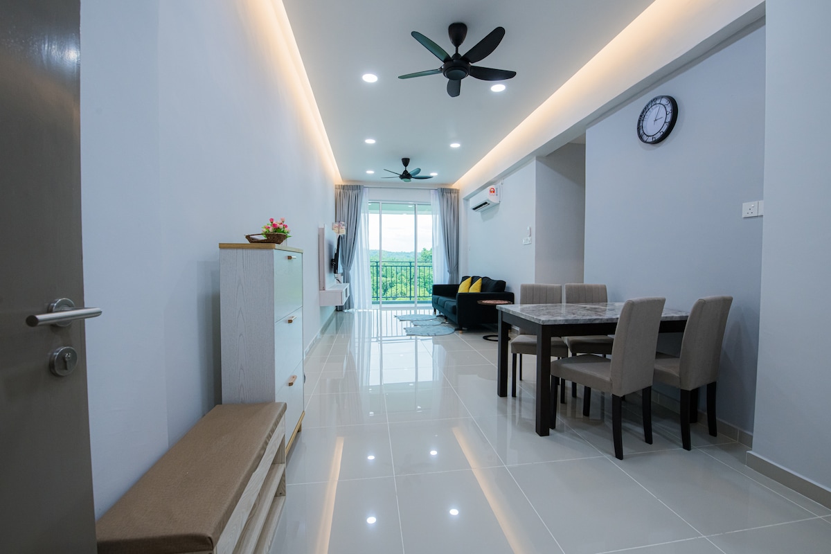 A bright and airy living space features large windows providing natural light and a view of greenery. A dining table is set for six, accompanied by gray upholstered chairs. A comfortable sofa in a contrasting color and decorative wall clock complete the serene atmosphere.