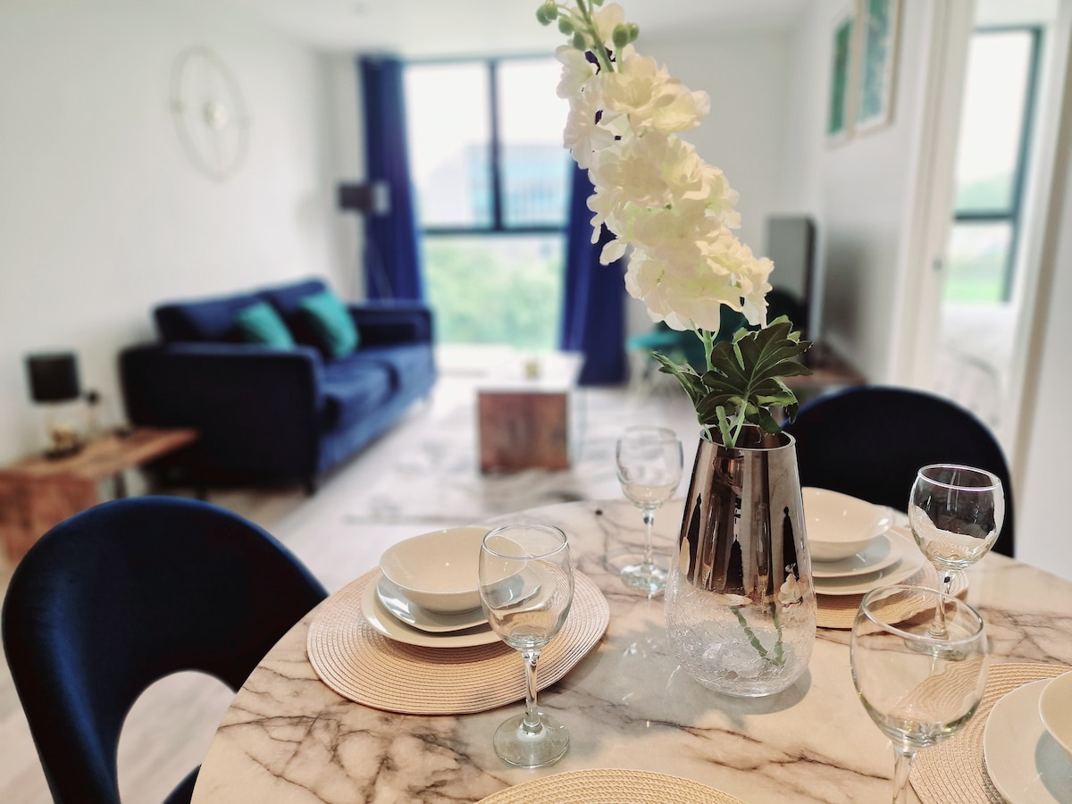 A modern dining area features a round marble table set for four, with clear glassware and neutral dinnerware. A decorative vase with white flowers stands at the center. In the background, a comfortable seating area with a dark blue sofa is visible, accented by cushions.