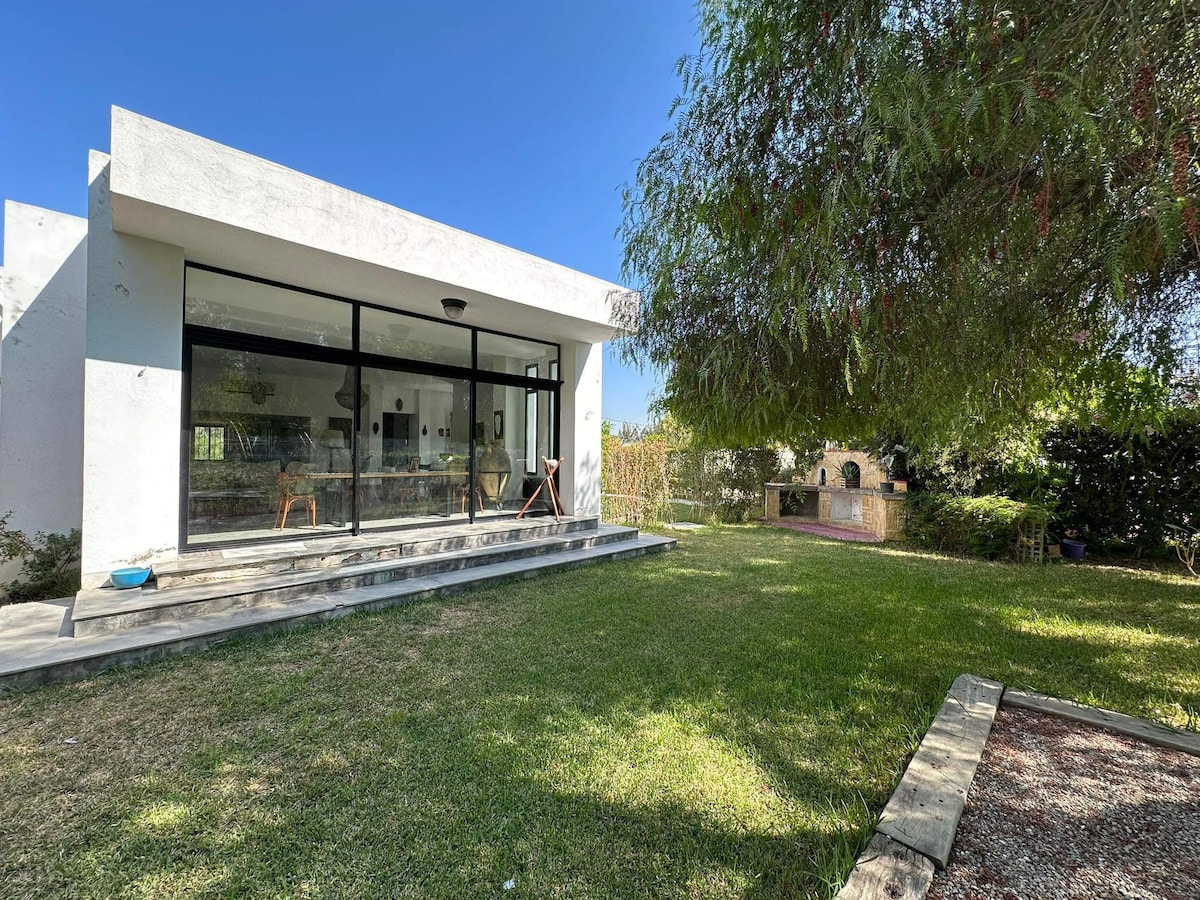 A modern villa is shown with a spacious terrace and large glass doors that provide natural light. Lush green grass surrounds the property, complimented by trees and a distant outdoor seating area. A peaceful atmosphere is created by the minimalist landscaping.
