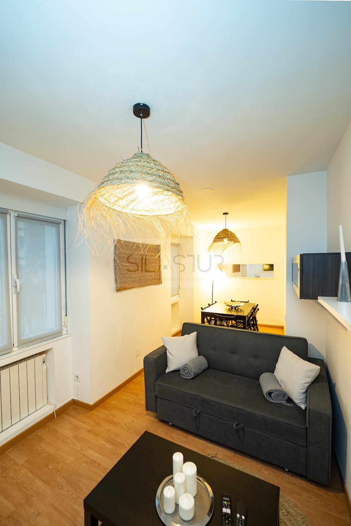 A cozy living area is presented with a dark-colored sofa, accented by light-colored cushions. Subtle lighting is provided by two pendant lights, enhancing the space's atmosphere. A glass-top table with candles sits in front of the sofa, while a dining area is visible in the background.