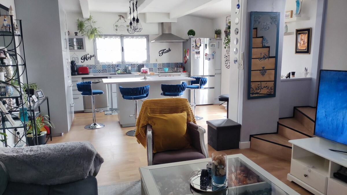 An open-concept living space is highlighted, featuring a modern kitchen with bar stools and a large countertop. Bright natural light fills the room through multiple windows, while plants and decorative items provide a lively touch. A comfortable seating area is visible with a cozy blanket draped on a chair.