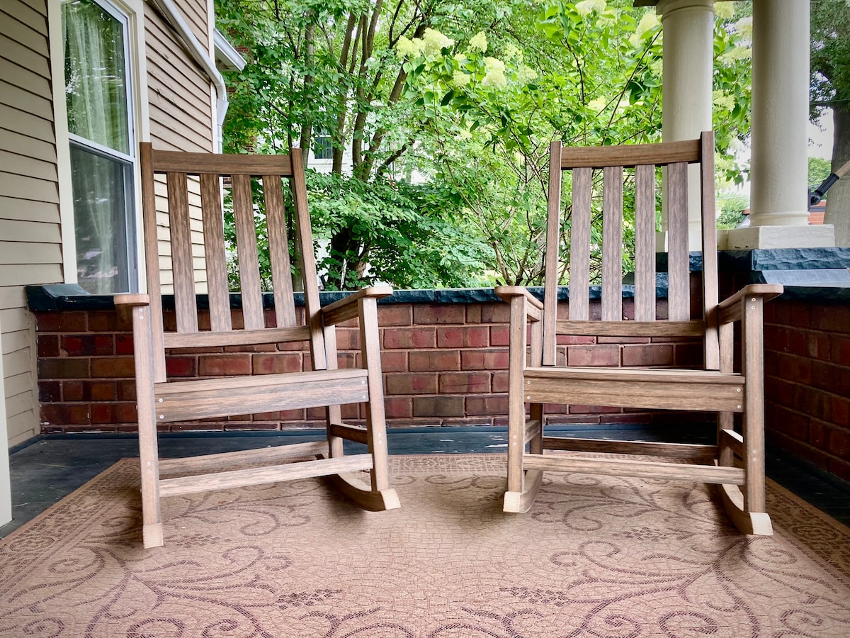 Two wooden rocking chairs are positioned on a porch, facing outward. A patterned area rug lies beneath them, and greenery is visible in the background, providing a natural and calming setting. The surrounding brick wall adds texture to the inviting outdoor space.