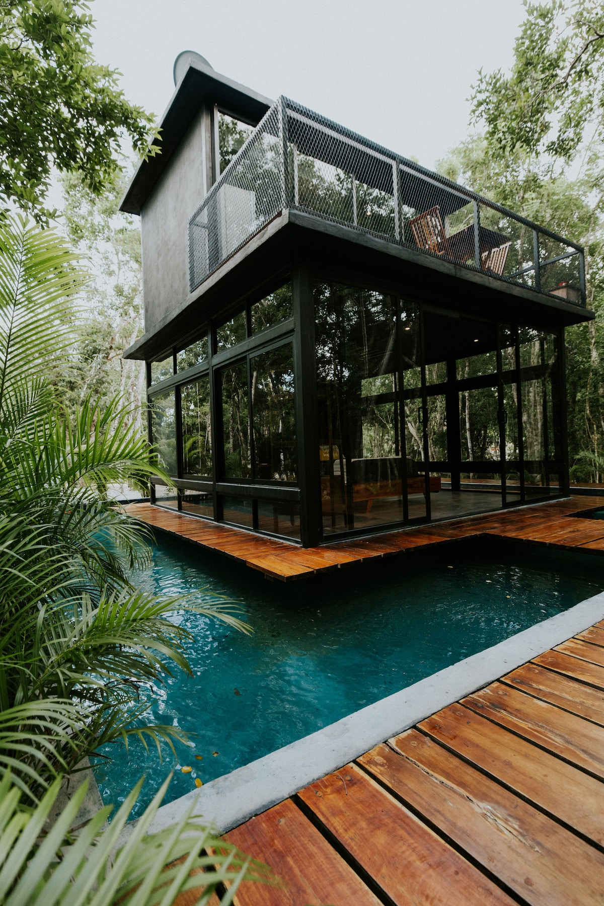 A modern structure elevated above water, featuring large glass walls that provide views of the surrounding greenery. The wooden deck leads to a serene pool, surrounded by lush foliage, creating a tranquil atmosphere.
