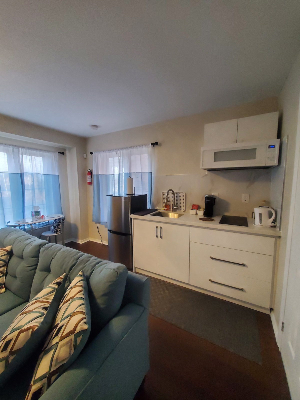 A cozy kitchenette is visible, featuring a refrigerator, microwave, and sink. A coffee maker and electric kettle are positioned on the counter. Natural light filters through sheer curtains, illuminating the room. A comfortable sofa is situated nearby, adorned with patterned cushions.