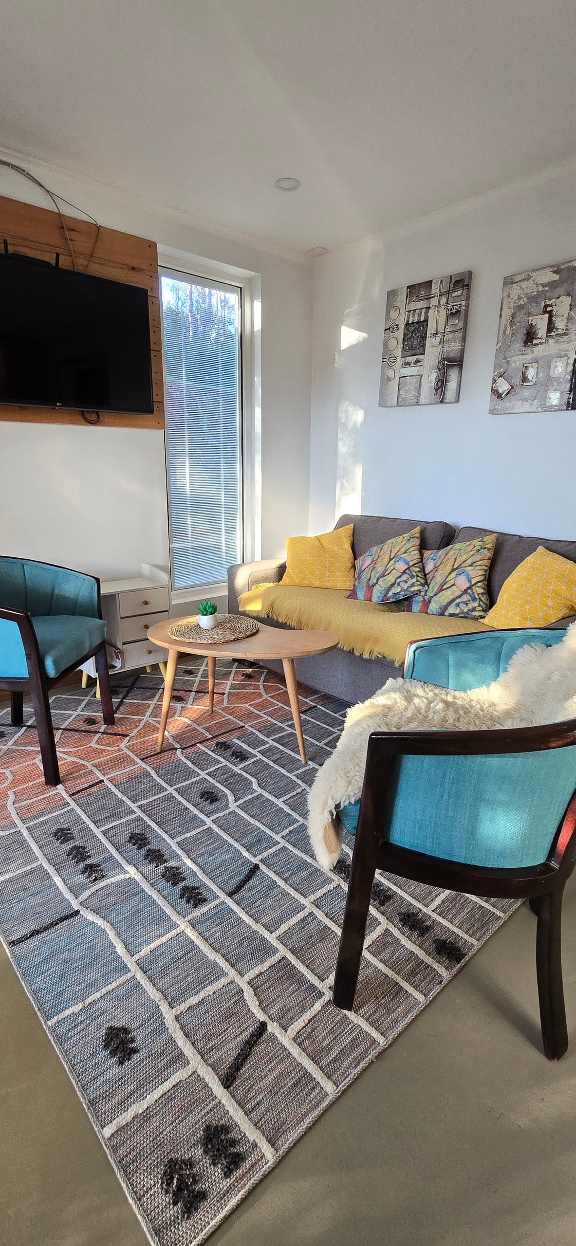 A comfortable seating area is highlighted by a gray sofa adorned with colorful cushions. Two blue chairs flank a small table, and a patterned rug anchors the space. Large windows allow natural light to brighten the room, complemented by minimalistic decor on the walls.