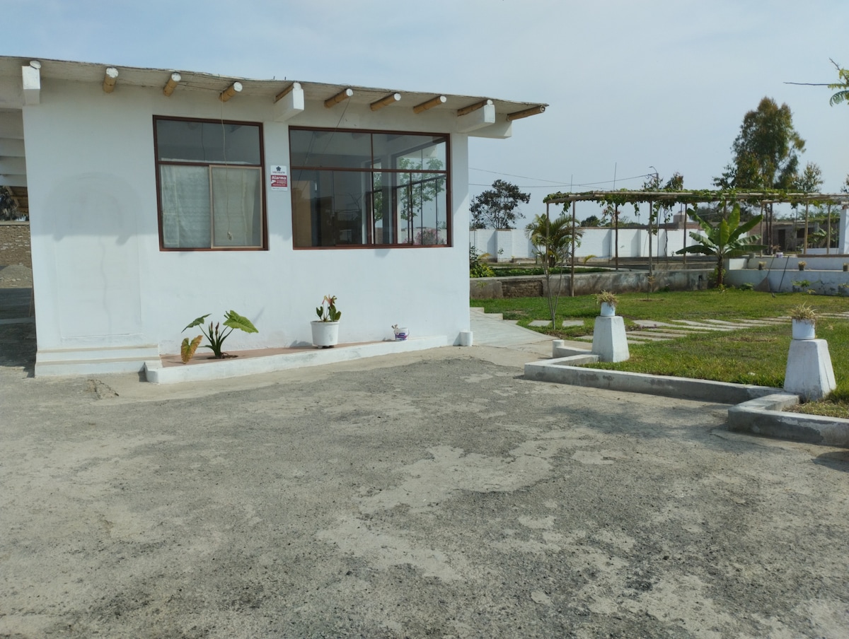 A white house with large windows is set against a backdrop of greenery. Potted plants are arranged near the entrance. The gravel surface allows for parking, and a spacious outdoor area features well-maintained grass and pathways, inviting relaxation and exploration.