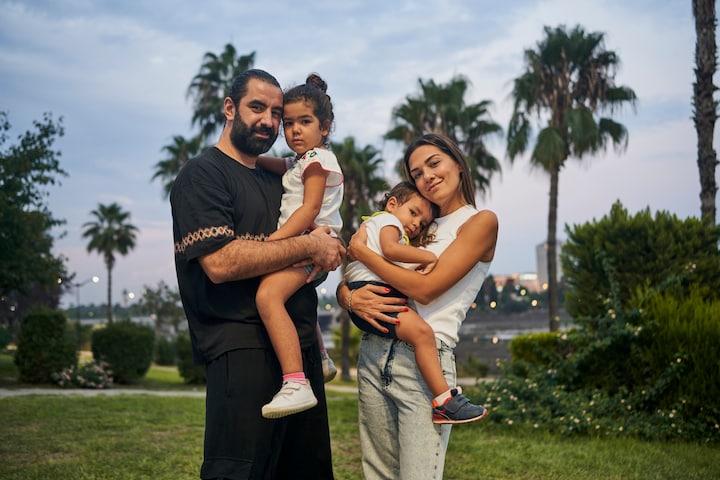 Un père et une mère se tiennent debout dans un parc avec des palmiers, leurs deux enfants dans les bras, et affichent un sourire paisible.