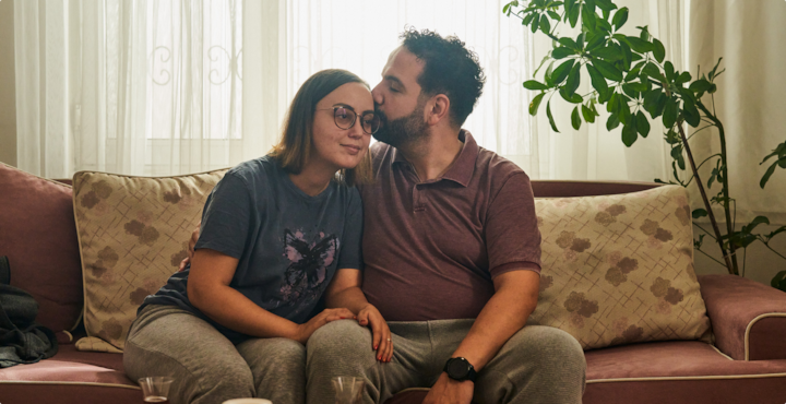 Two people sit on a couch, leaning into each other. Sunlight filters through sheer curtains behind them.